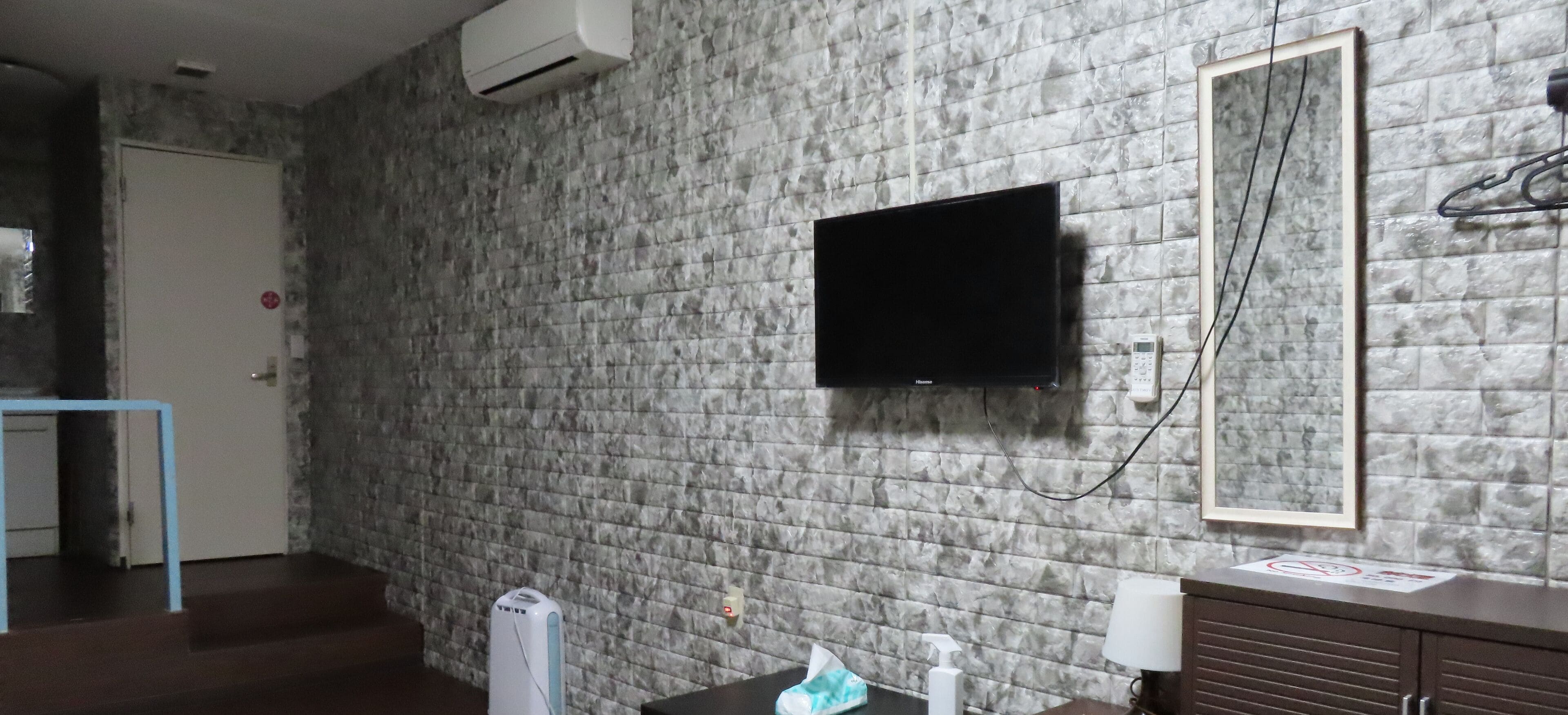 A bathroom with a stone-textured wall, a flat-screen TV mounted above a mirror, a towel dispenser, and a cabinet with a no-smoking sign.