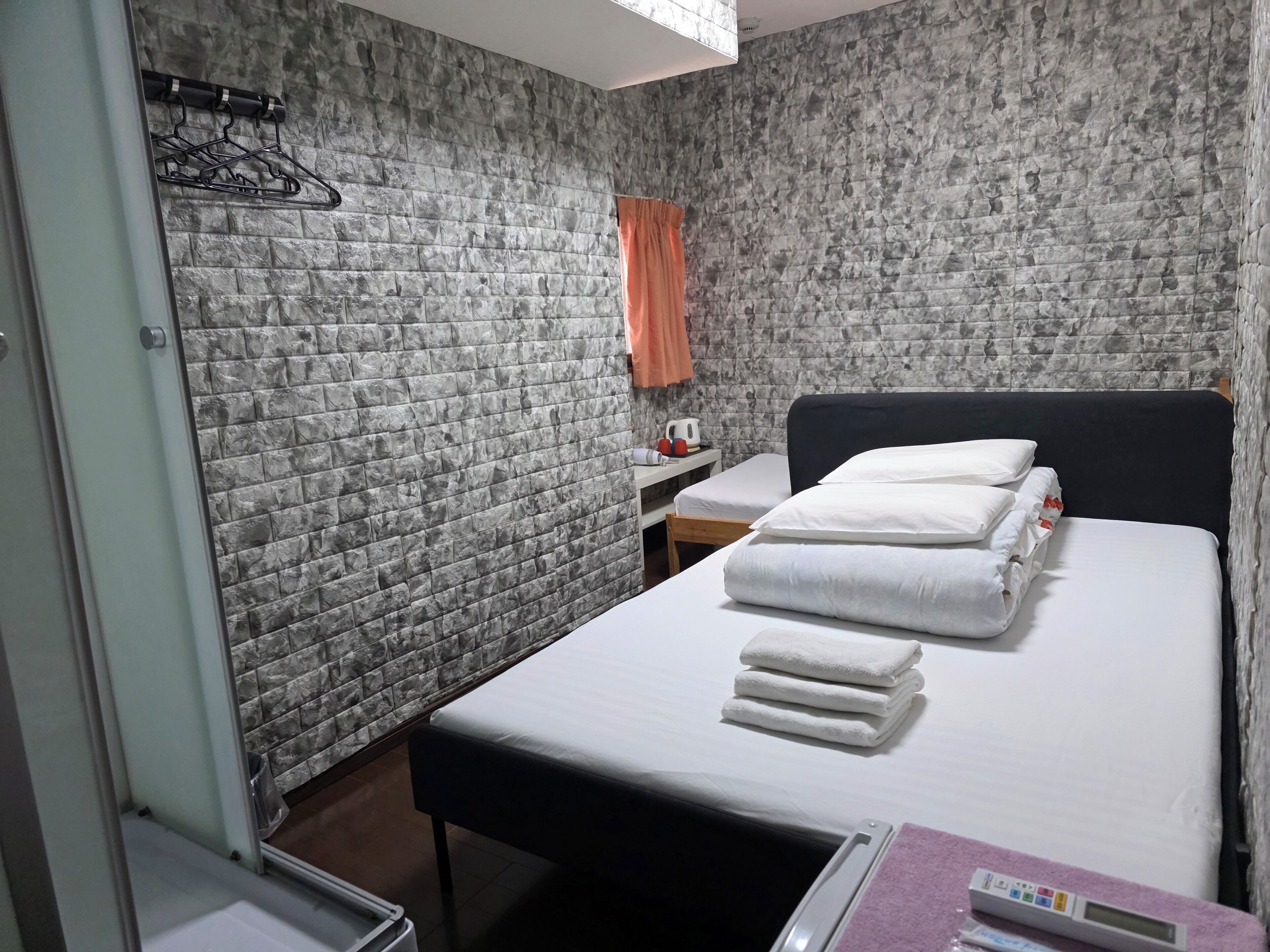 A hotel room with a bed, a small table with a kettle, a wall-mounted heater, and a towel rack.