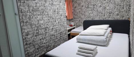 A hotel room with a bed, a small table with a kettle, a wall-mounted heater, and a towel rack.