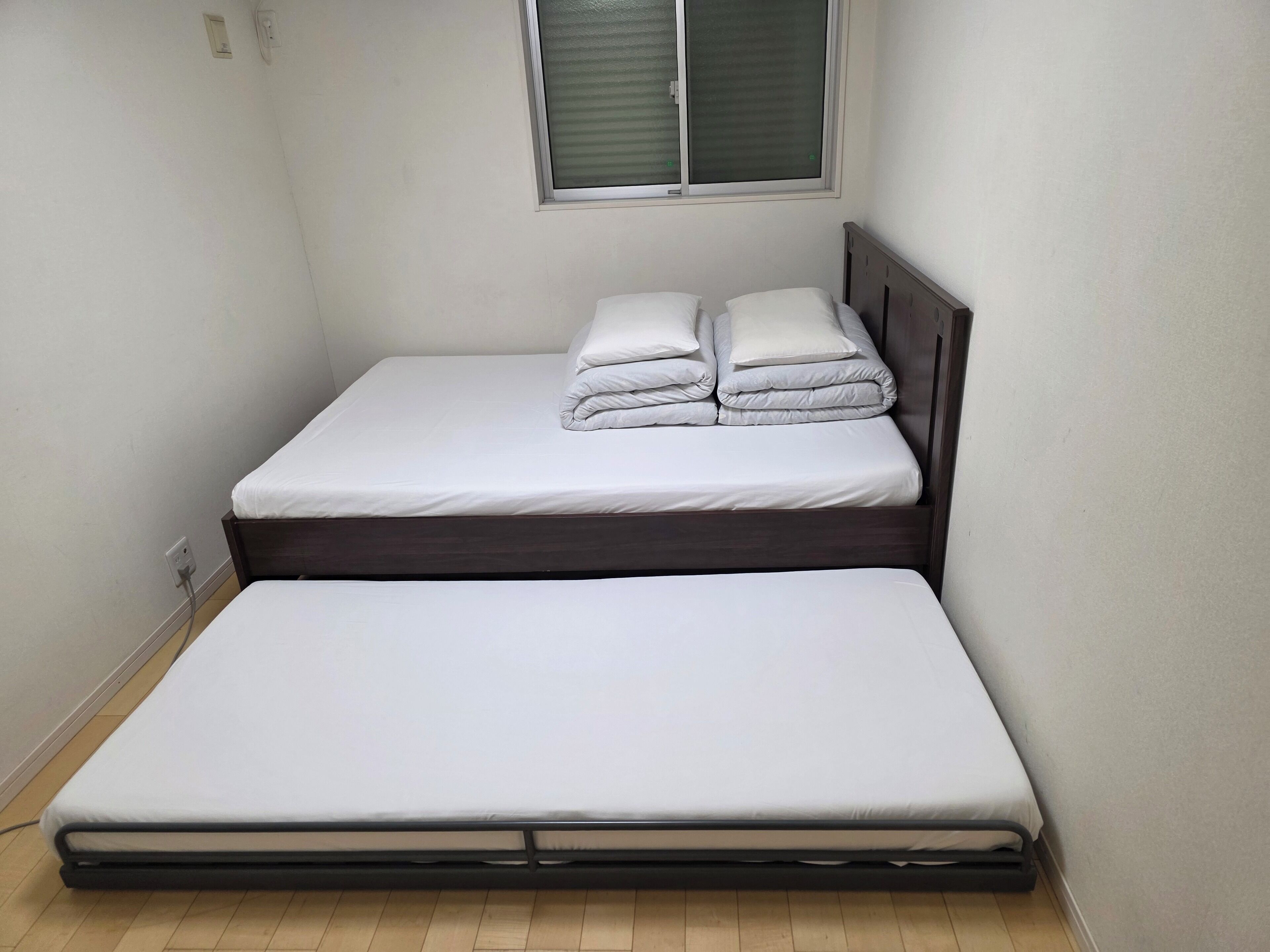 A small, single-bed room with a wooden headboard, a window with blinds, and a power outlet on the wall.