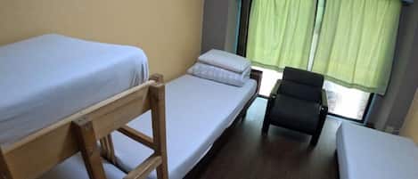 Habitación pequeña y funcional con dos camas individuales, una escalera de madera, una silla y una ventana con cortinas verdes.