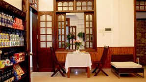 A room with a wooden table, two chairs, a small bench, and a shelf with bottled water and snacks. - Green Grass Homestay (Hoi An)