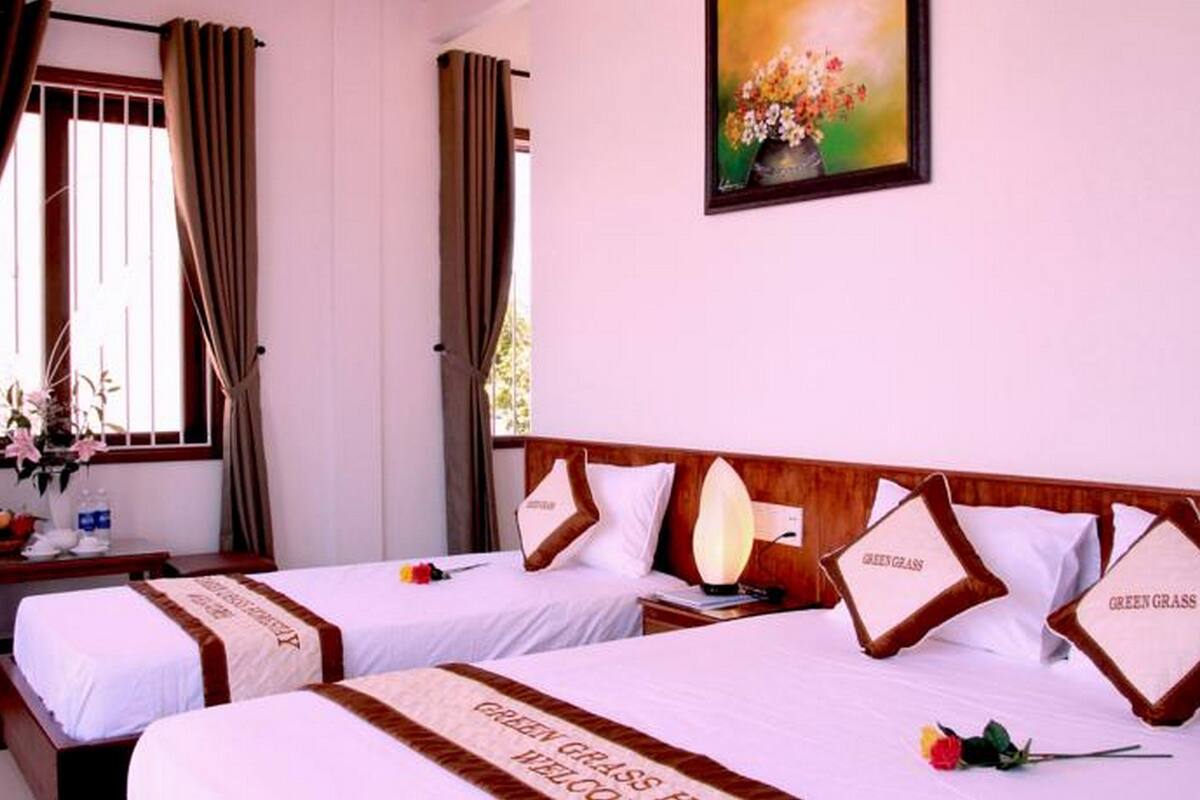 A hotel room with two beds, each with a 'Green Grass' bedspread, a bedside table with a lamp, a vase of flowers, and a painting on the wall.