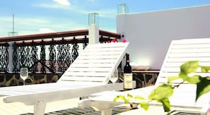 A rooftop terrace with white lounge chairs, a bottle of wine, and a glass of wine on a table.