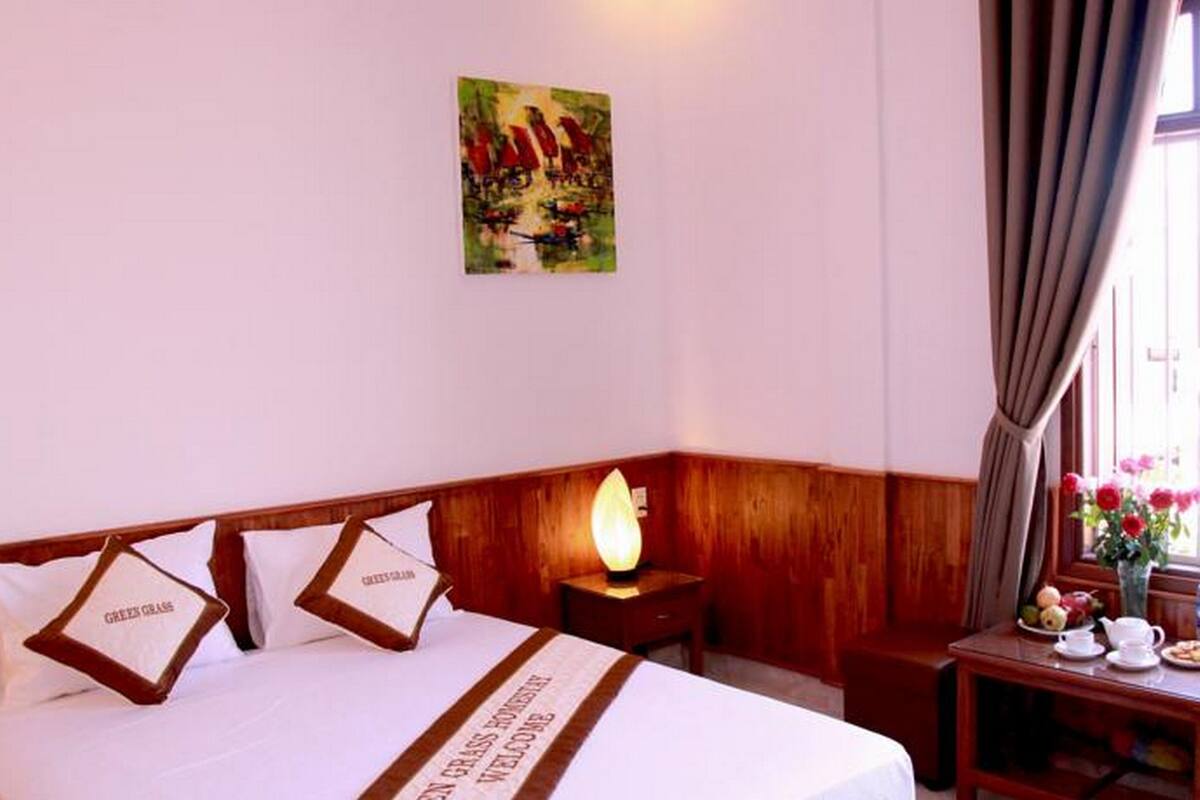 A hotel room with a bed, a nightstand with a lamp, a small table with a vase of flowers, and a window with curtains.