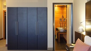 A hotel room with a large wardrobe, a bathroom with a sink and mirror, and a bedside table with a lamp. - Tana Hôtel (Antananarivo)