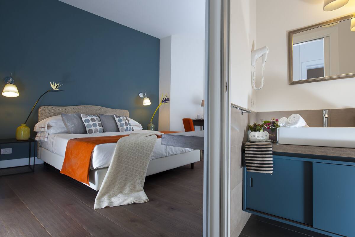 A modern bedroom with a bed, bedside lamps, a nightstand, and a bathroom with a sink and mirror.