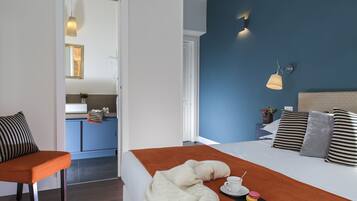 Une chambre d’hôtel avec un lit, une chaise, une petite table avec une tasse et une pâtisserie, et une vue sur la salle de bain et la cuisine.
