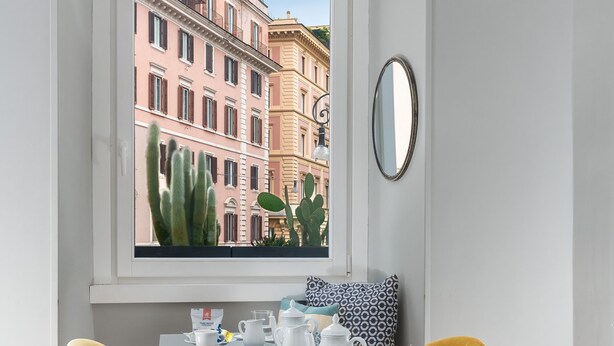 Un coin repas avec une table dressée pour quatre personnes, agrémenté de chaises jaunes et d’une table en verre. Une fenêtre offre une vue sur des immeubles et un cactus.