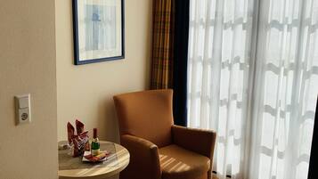 Quarto de hotel com poltrona marrom, uma mesinha redonda com petiscos e uma garrafa, um quadro na parede e uma janela grande com cortinas leves.