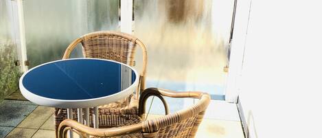 A patio area with a round table and two wicker chairs, a glass partition, and a view of a residential building.