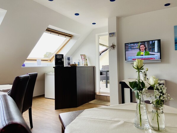 A modern hotel room with a dining area, a kitchen, and a TV displaying a news channel.