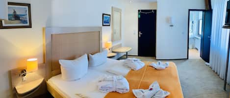 Habitación de hotel con cama, mesita de noche, lámpara, perchero para toallas y una puerta.