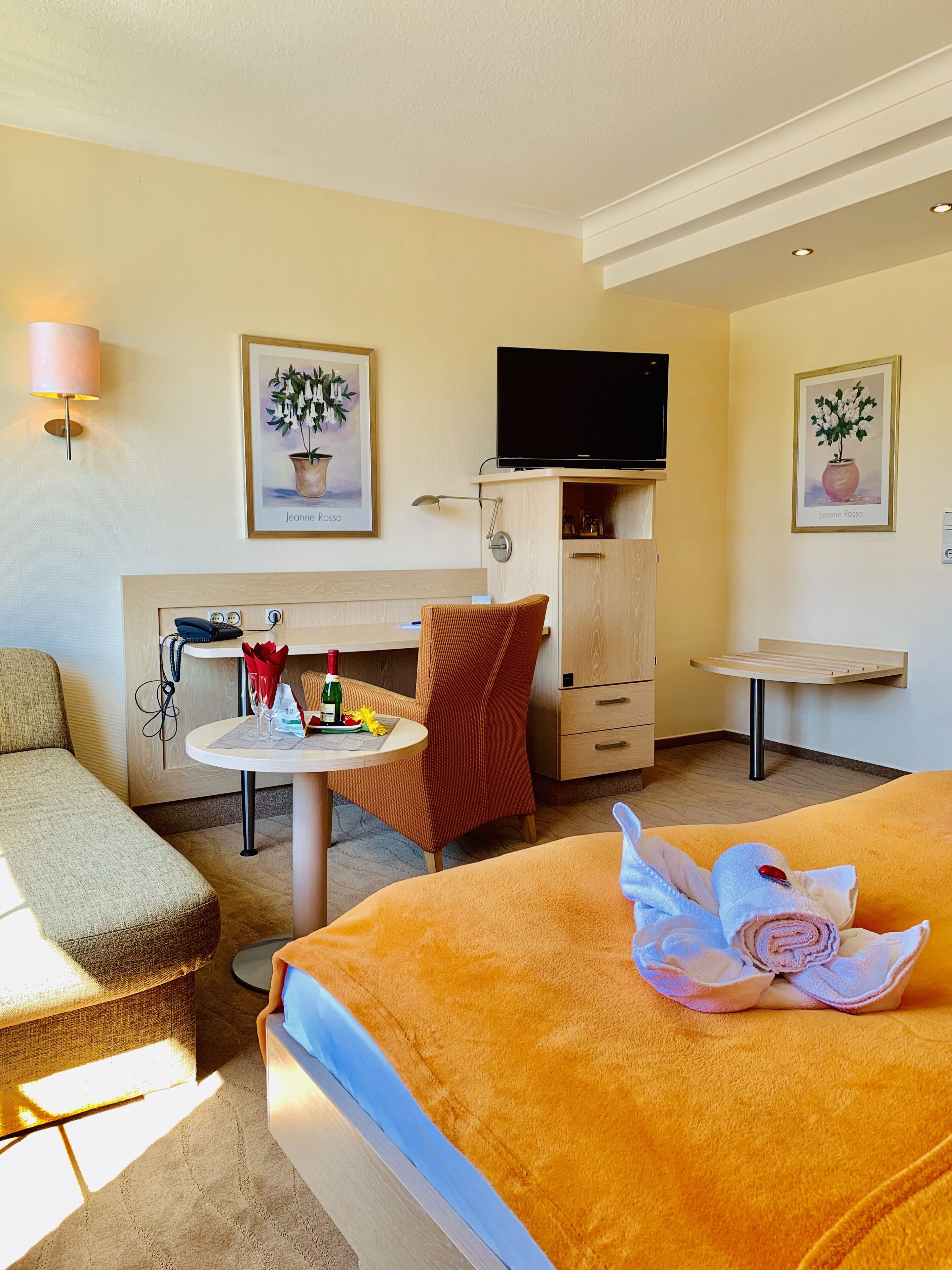 A hotel room with a bed, a desk, a chair, a TV, and artwork on the walls.