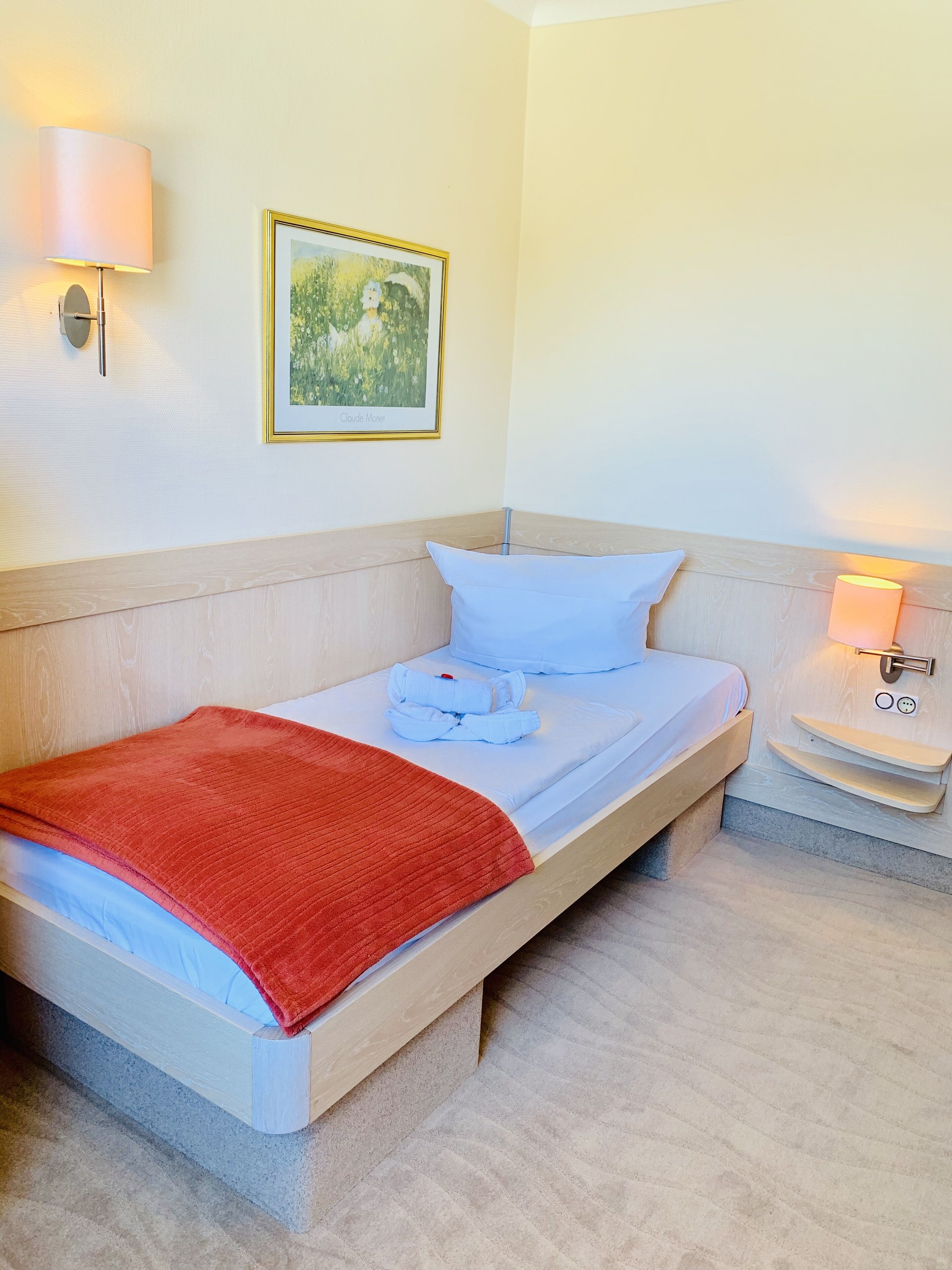 A neatly made bed with a red blanket, a white pillow, and a folded white towel. There is a framed picture on the wall above the bed and a wall-mounted lamp beside the bed.