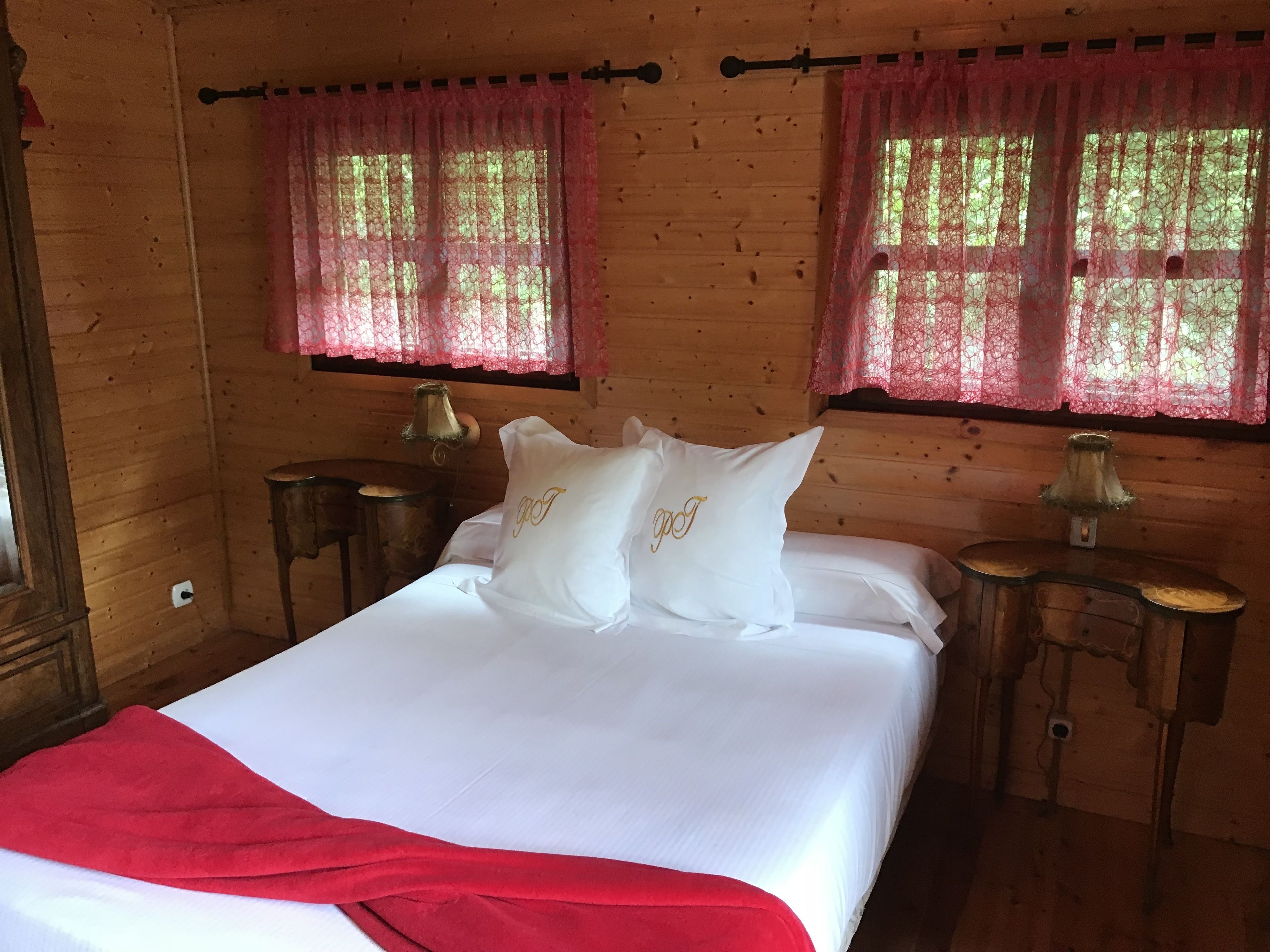 A wooden cabin room with a bed, two nightstands, and two windows with lace curtains.
