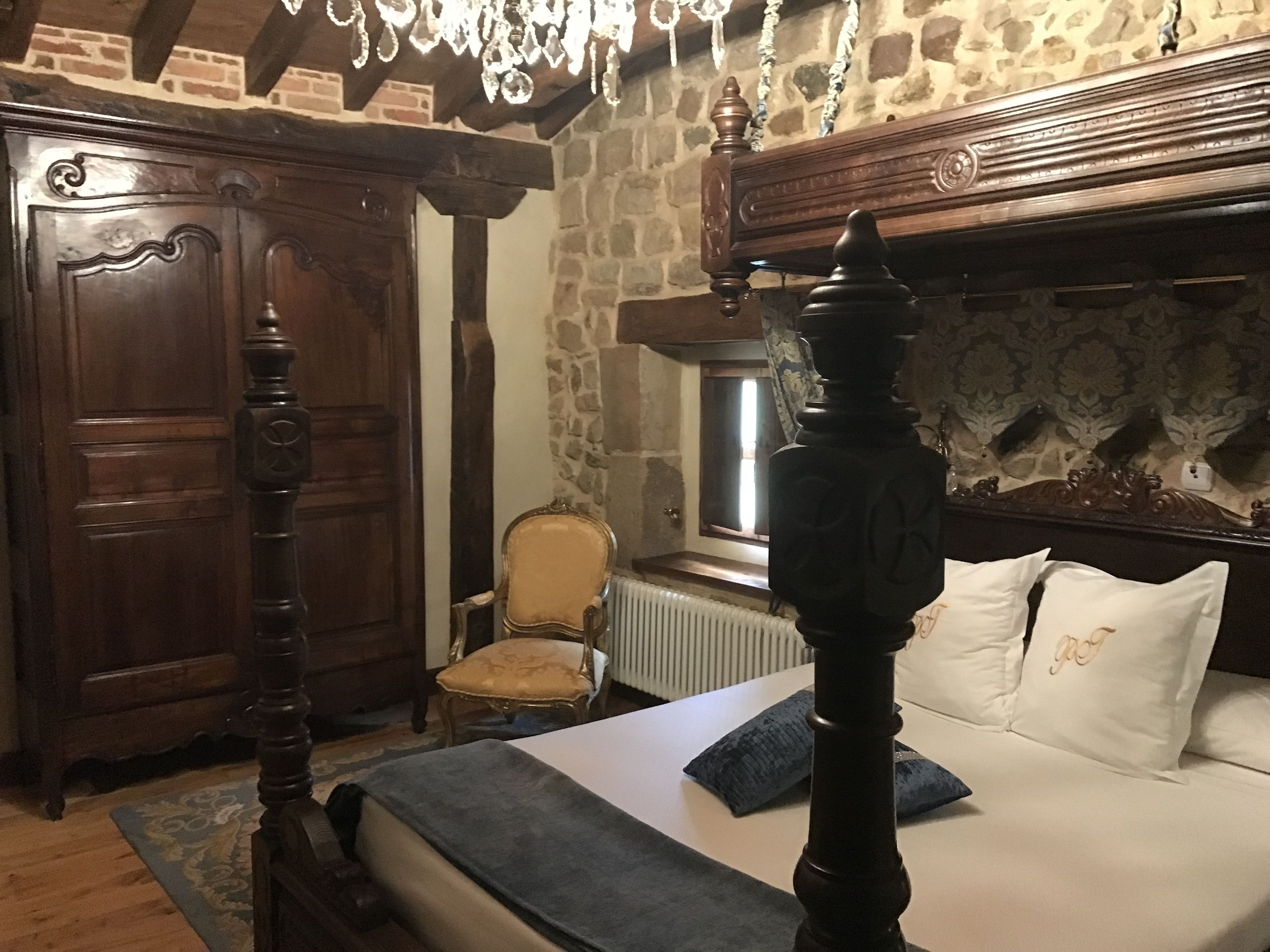 A bedroom with a four-poster bed, a wooden wardrobe, a chair, and a stone wall.