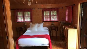 Un dormitorio de cabaña de madera con una cama, dos mesitas de noche, un candelabro y ventanas con cortinas rojas.