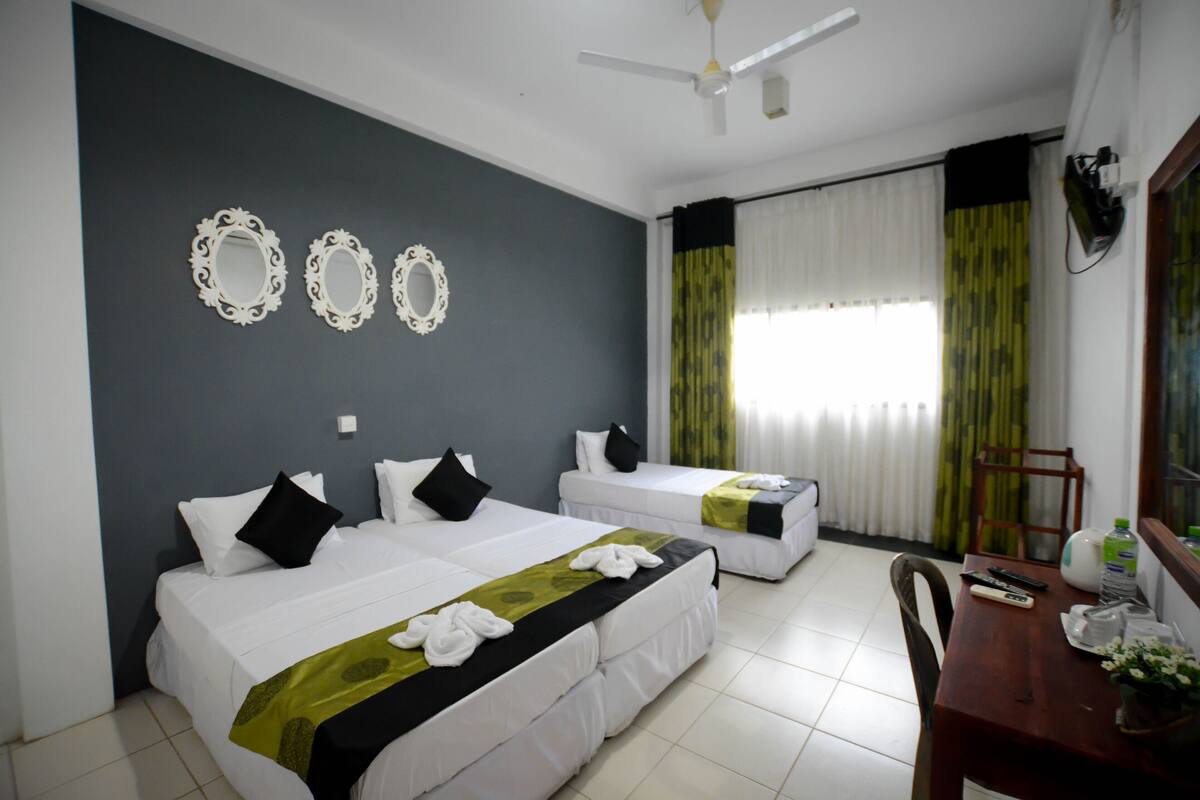 A hotel room with two beds, a dining table, a ceiling fan, and a window with curtains.
