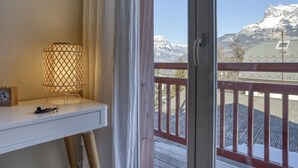 Ein Zimmer mit weißem Schreibtisch, einer Lampe und Blick auf schneebedeckte Berge durch eine Balkontür.