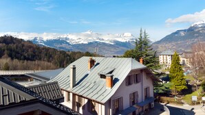 Ein Gebäude mit schrägem Dach und mehreren Schornsteinen, eingebettet in eine gebirgige Landschaft mit schneebedeckten Gipfeln.
