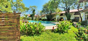 A pool surrounded by lush greenery, with a wooden fence and a building in the background.