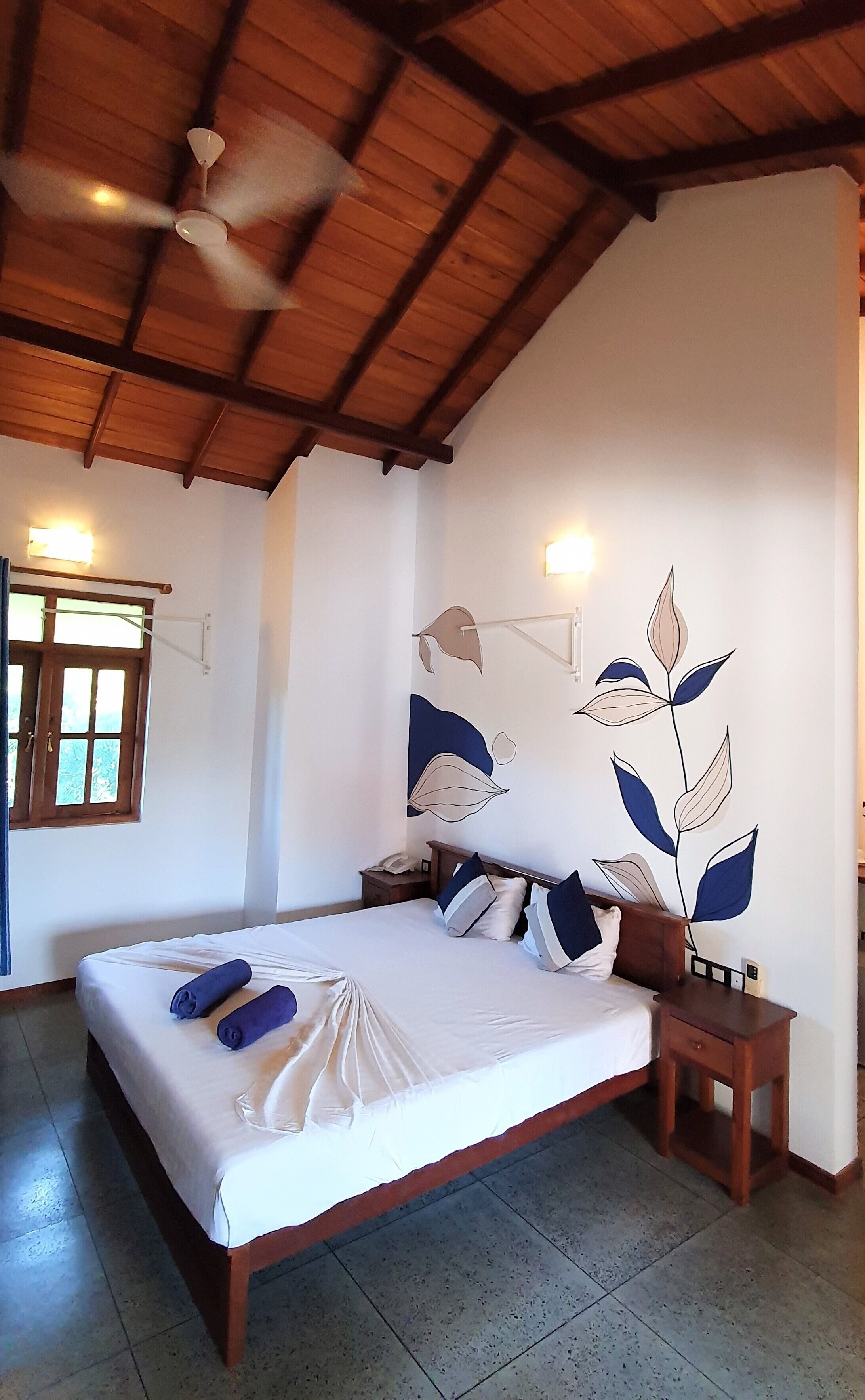 A bedroom with a large bed, wooden headboard, bedside tables, a ceiling fan, and a wall with a leaf design.
