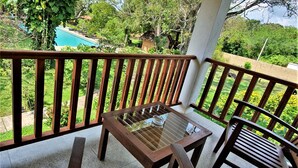 Un balcón de madera con mesa y sillas de vidrio, con vistas a una piscina y abundante vegetación.