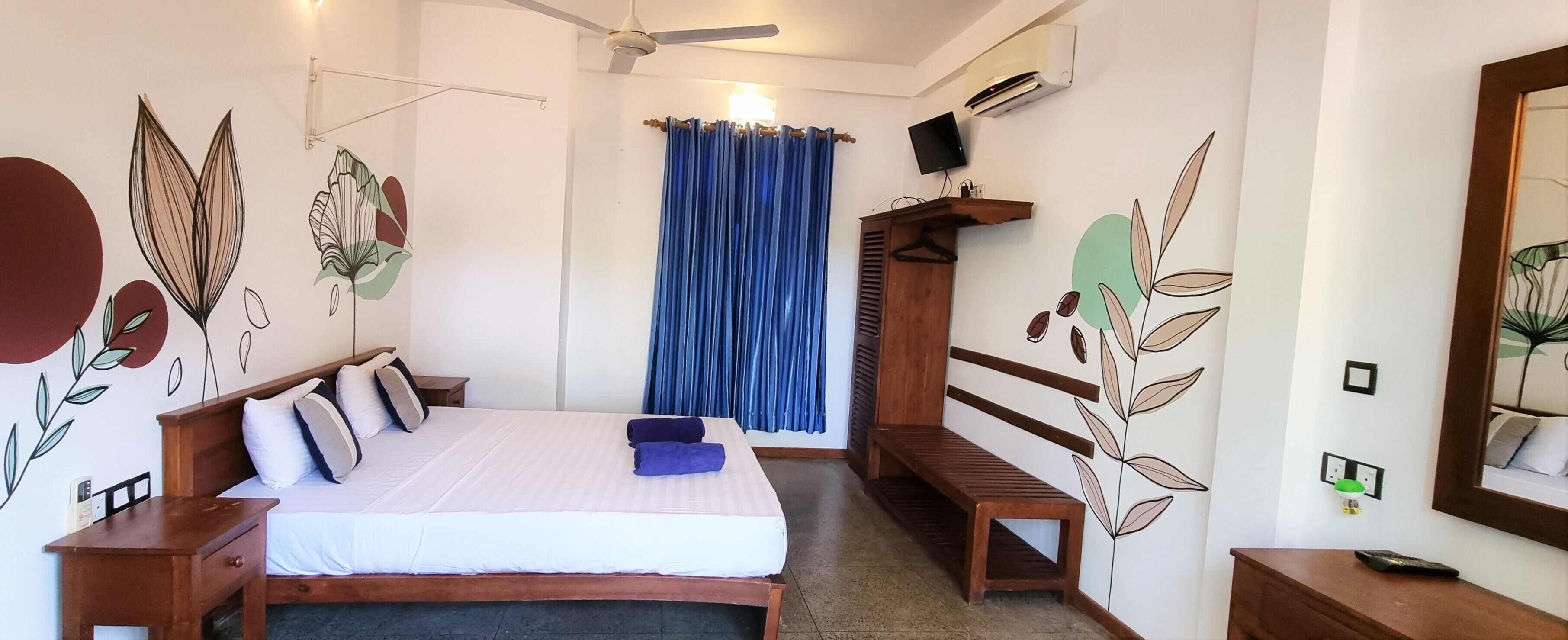 Un dormitorio con cama, mesitas de noche, un banco, ventilador de techo, televisor y ventana con cortinas.