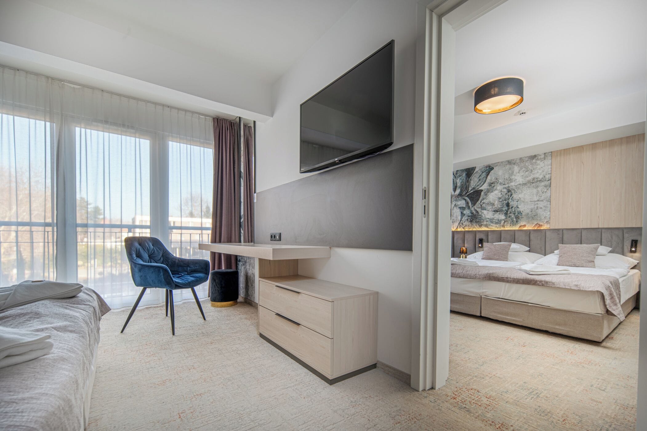 Ein modernes Hotelzimmer mit einem großen Bett, einem Schreibtisch mit Flachbildfernseher, einem Sessel und einem großen Fenster mit Vorhängen.