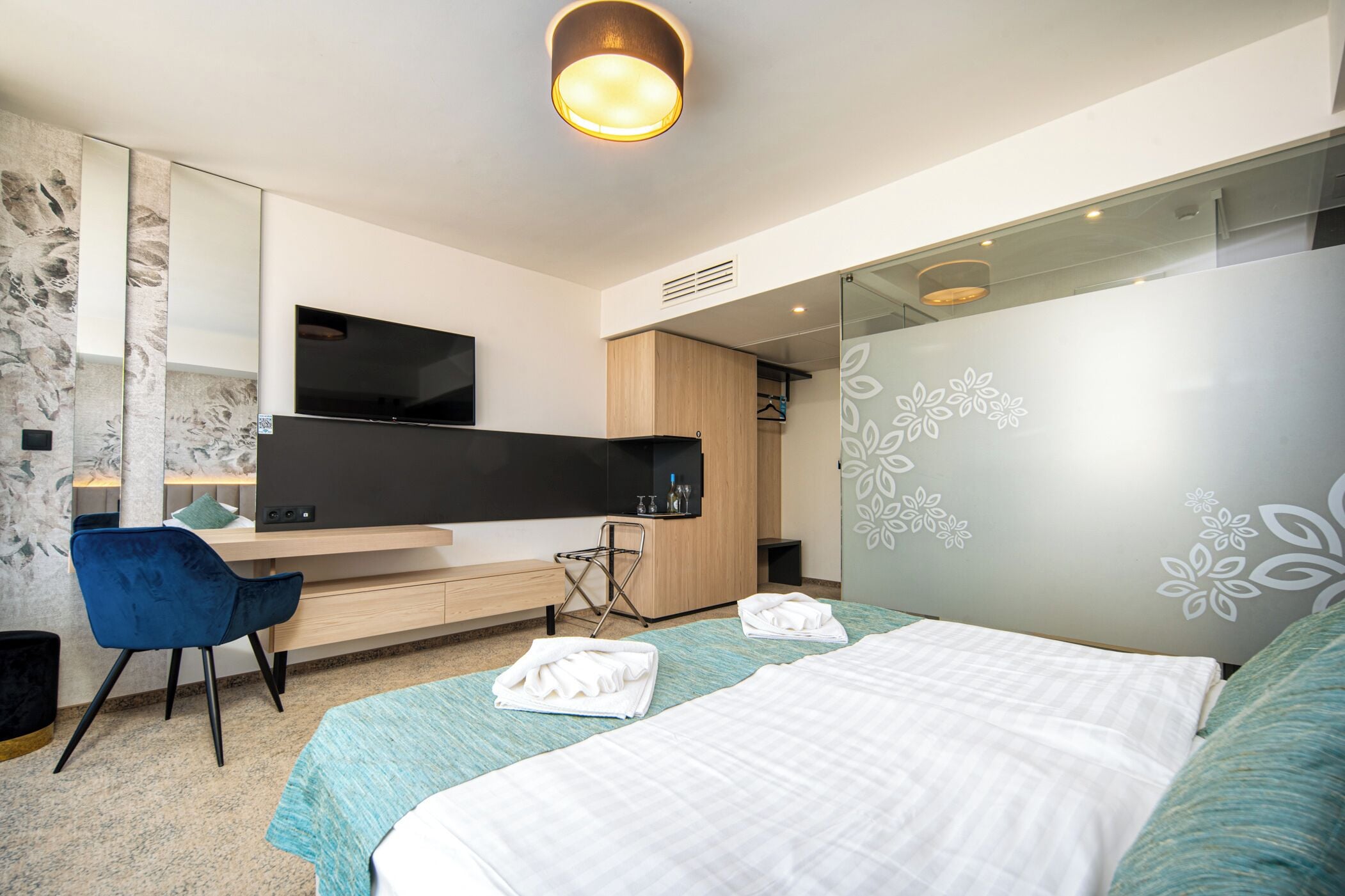 Une chambre d’hôtel moderne avec un grand lit, un bureau doté d’un téléviseur à écran plat, une chaise bleue et une armoire.