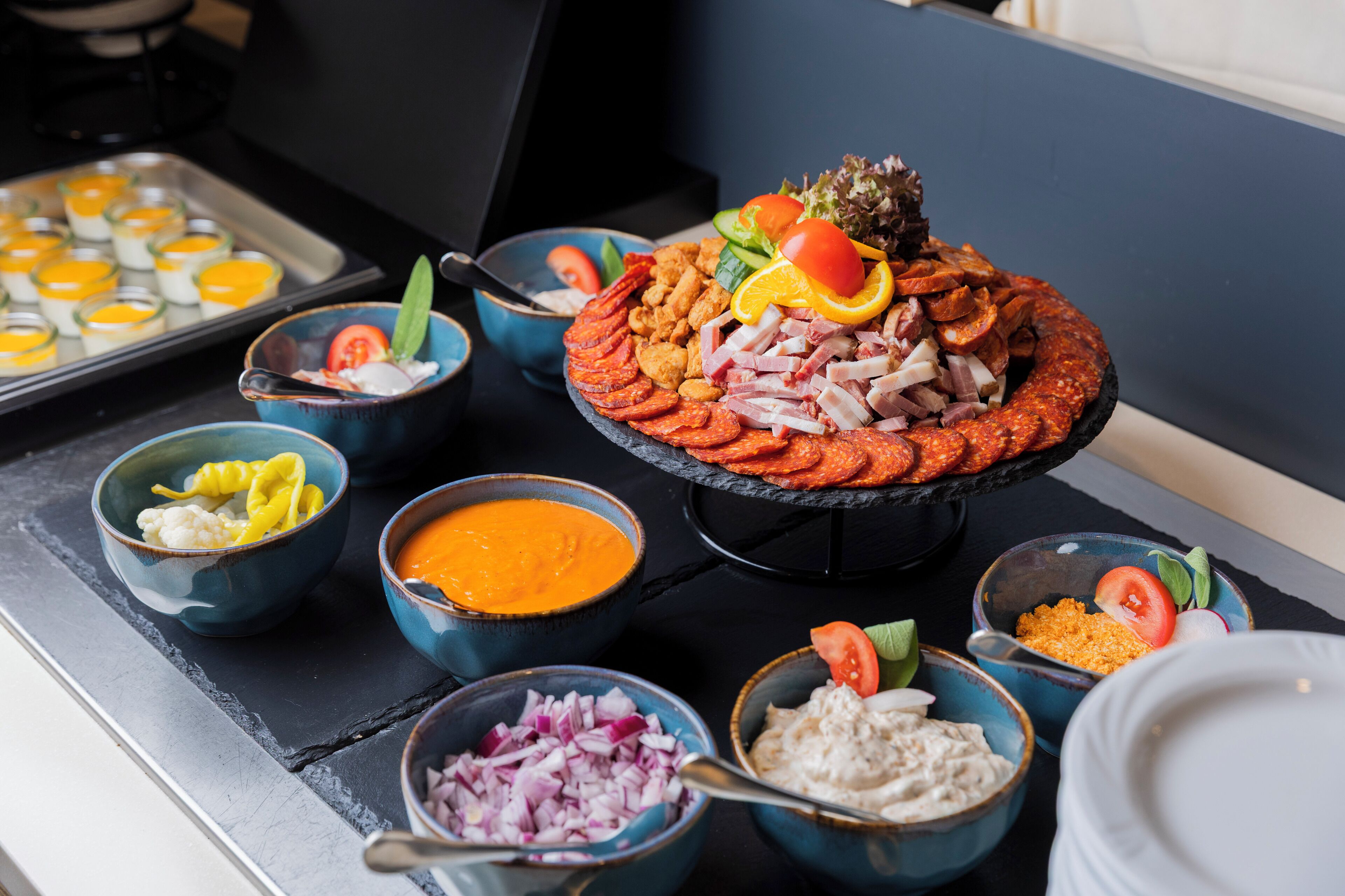 Diverse soorten eten, waaronder een groot schaal met vleesschijfjes, groenten en een kom gele eiercustard.