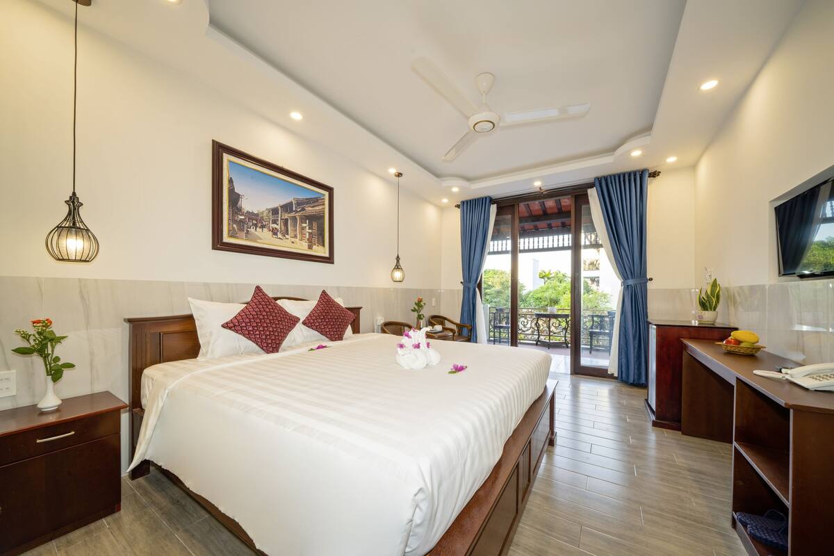 Une chambre d’hôtel avec un grand lit, un ventilateur de plafond, un bureau avec un téléphone et un balcon donnant sur un paysage verdoyant.