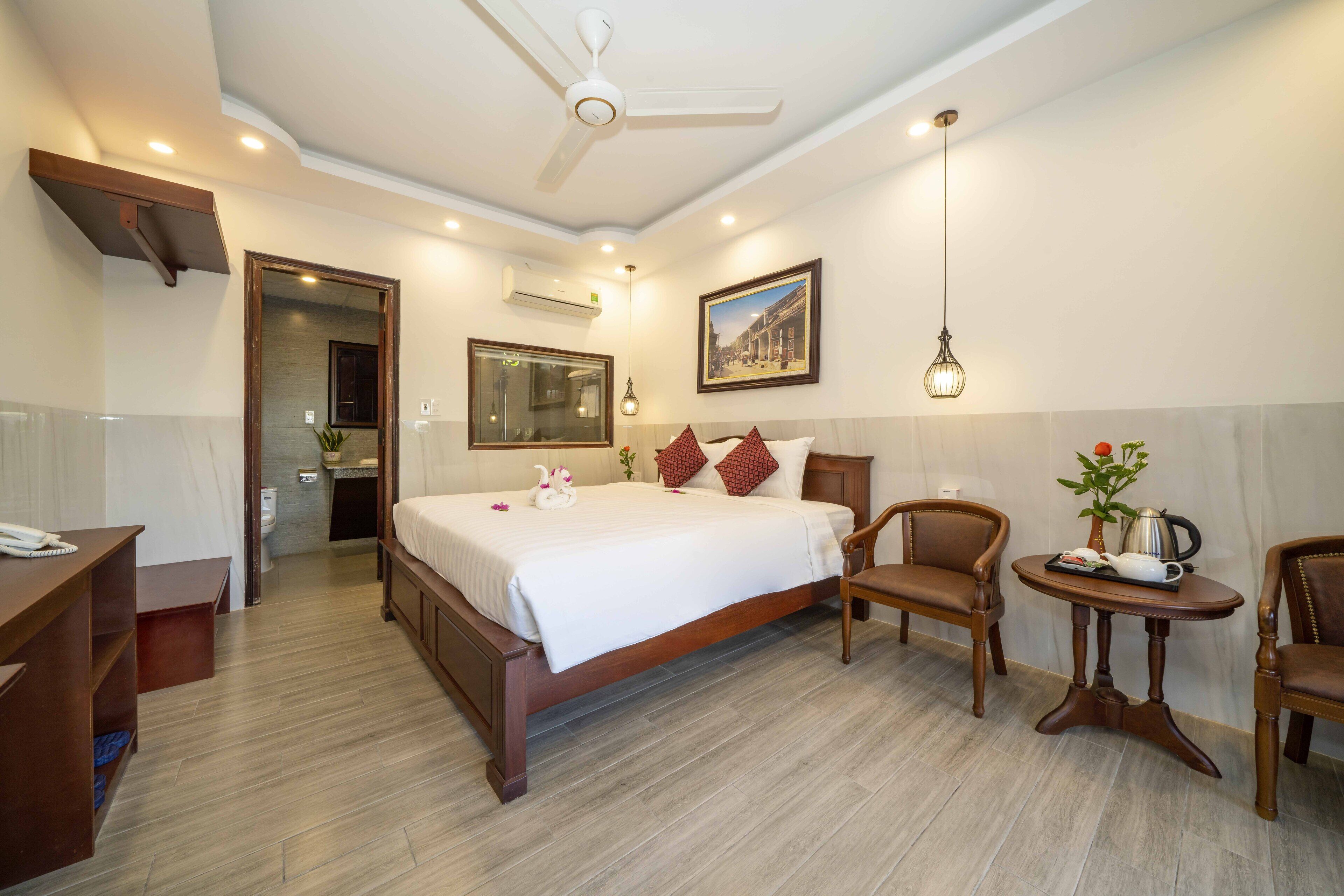 Une chambre d’hôtel avec un grand lit, des meubles en bois, une petite table avec un vase et un ventilateur de plafond.