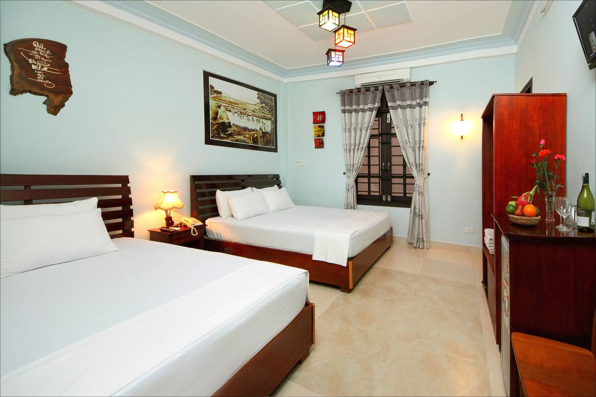 A hotel room with two beds, a wooden headboard, a nightstand, a lamp, a wall-mounted painting, a window with curtains, a wooden cabinet with fruit and a vase, and a ceiling fan.