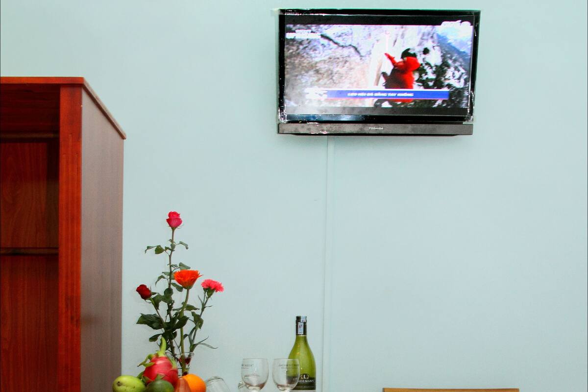 A small refrigerator with a Sanyo sticker, a wooden chair, a television mounted on the wall, and a table with a fruit bowl, wine glasses, and a bottle.