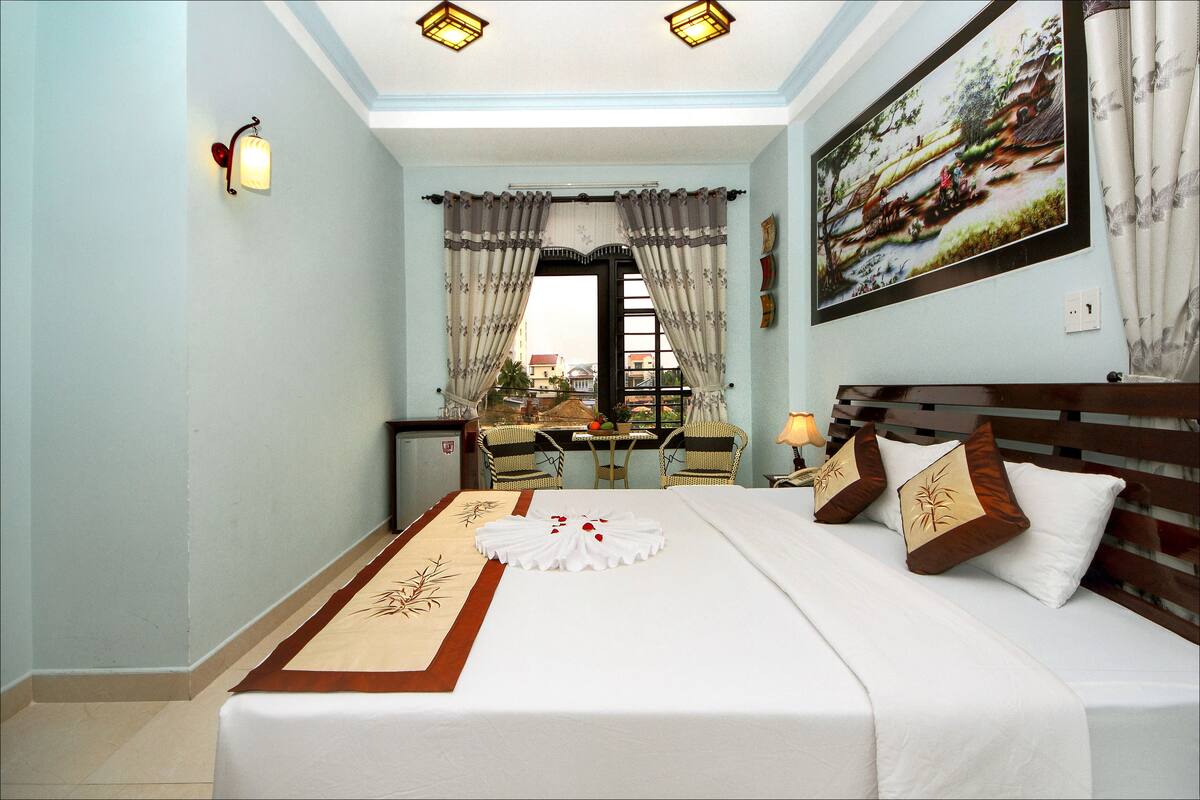A hotel room with a neatly made bed, a dining area with chairs and a table, and a large window with curtains.