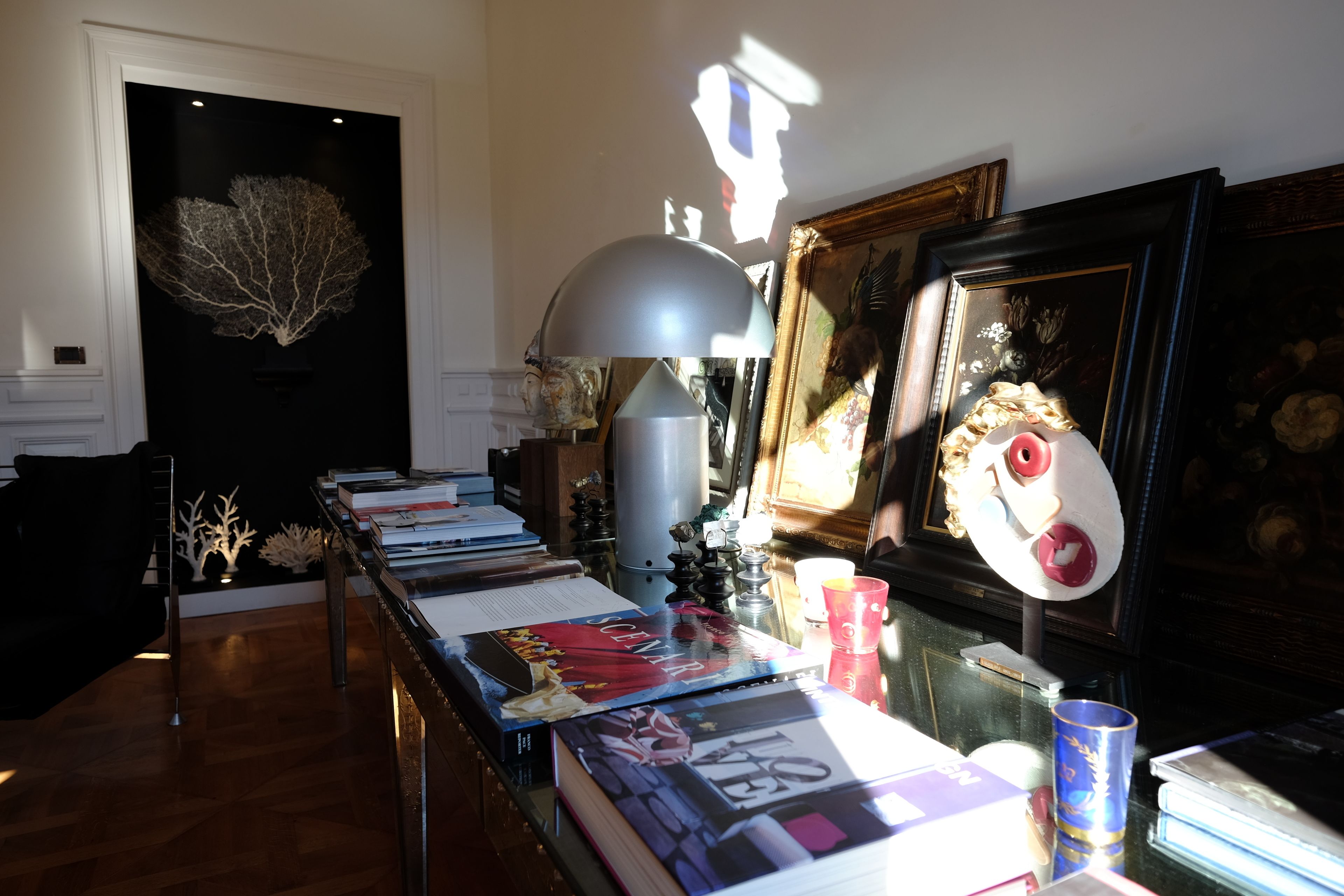 A room with a long table displaying books and decorative items, a lamp, and a framed artwork on the wall.