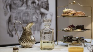 Une table avec un présentoir à gâteaux à trois niveaux, un pichet en argent, un verre de jus d’orange et, en arrière-plan, une œuvre d’art encadrée.