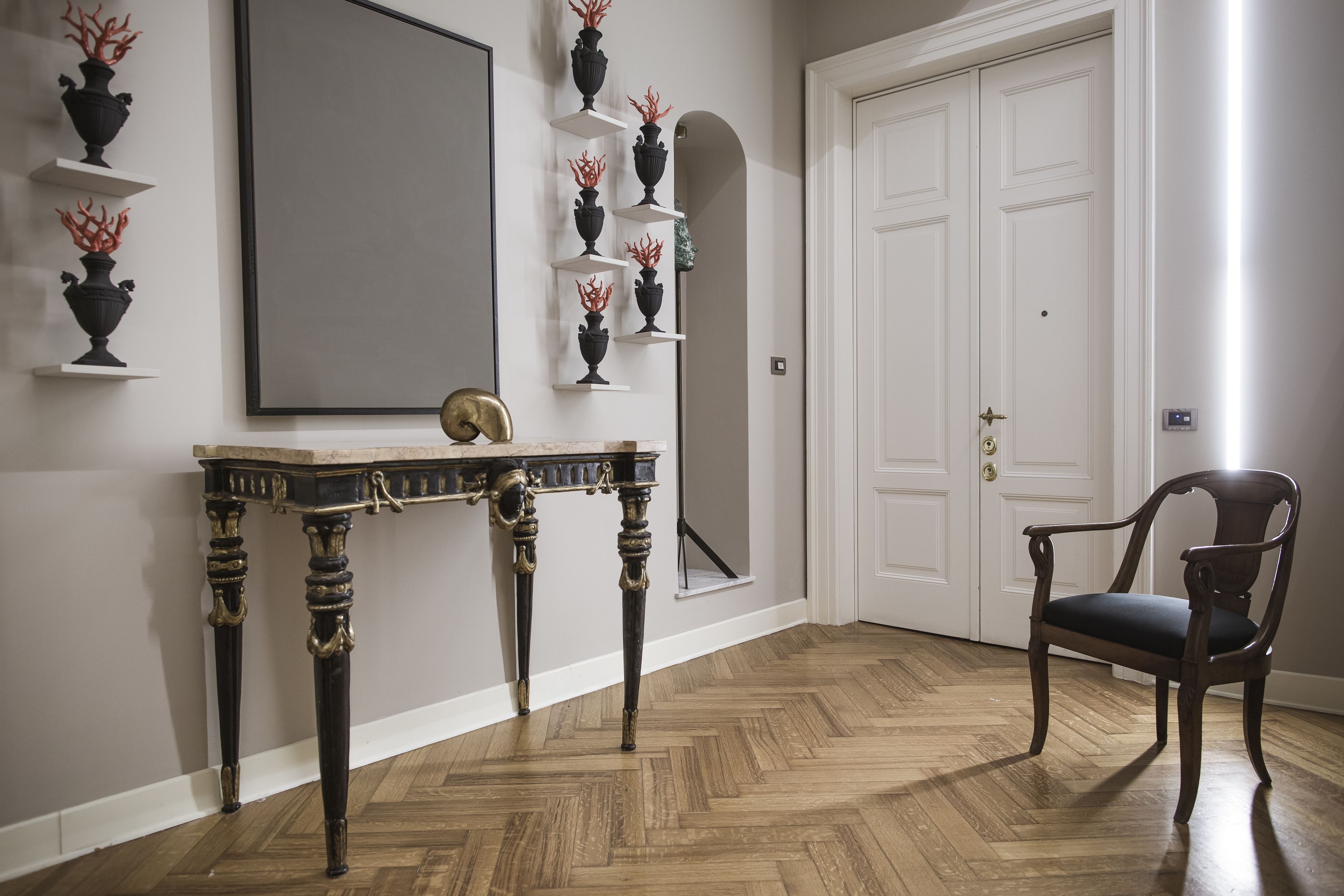 Un corridoio con pavimento in parquet a spina di pesce, un tavolino consolle in legno scuro con un oggetto dorato sopra, e una sedia in legno scuro.