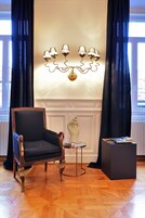 A room with a dark blue armchair, a small round table with books, a decorative sculpture, and a wall-mounted lamp.