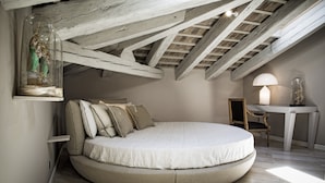 Un lit rond dans une chambre à coucher aménagée dans les combles, avec des poutres apparentes, une petite table avec une lampe et un objet décoratif accroché au mur.