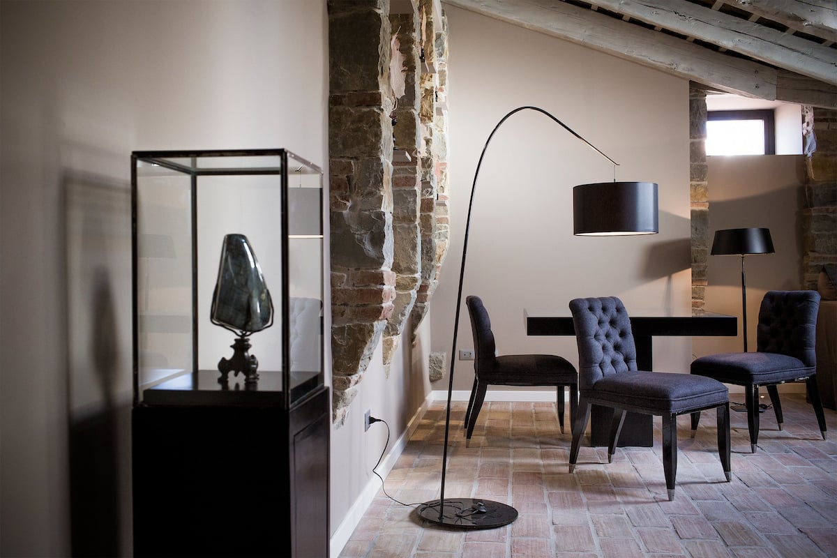 Quarto com parede de pedra, uma vitrine de vidro com luminária, duas cadeiras, um abajur e uma mesa.