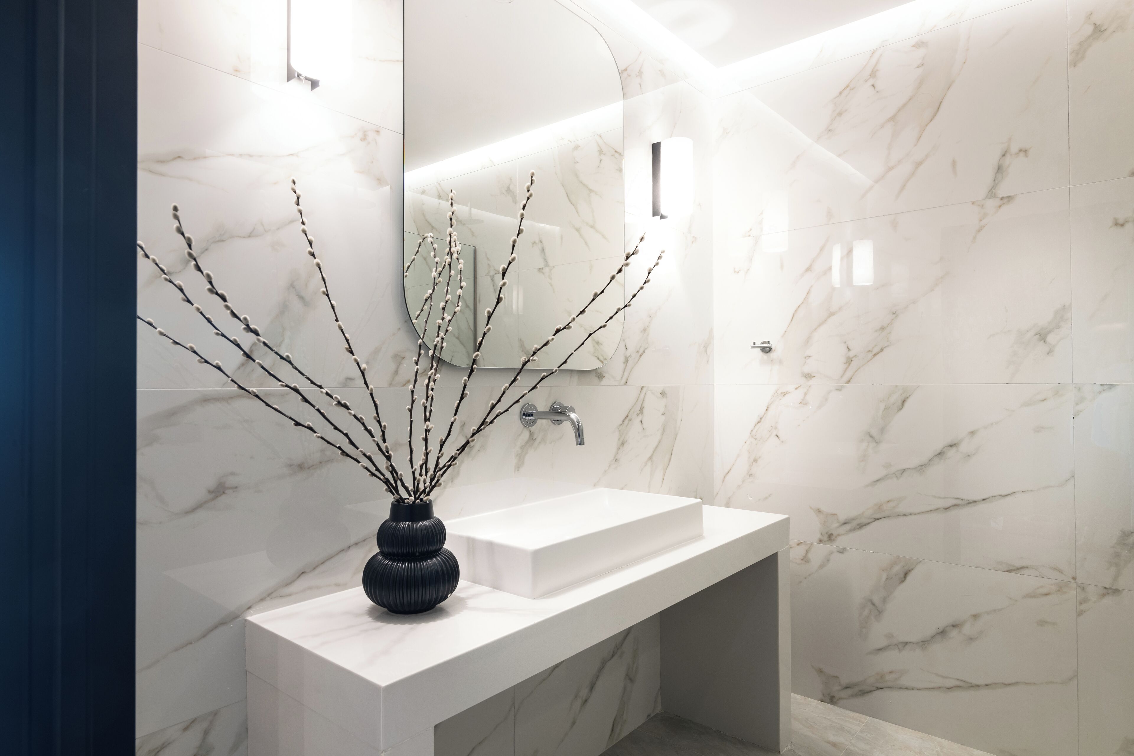 Un baño moderno con un lavamanos de mármol blanco, un jarrón negro con ramas y un espejo grande.