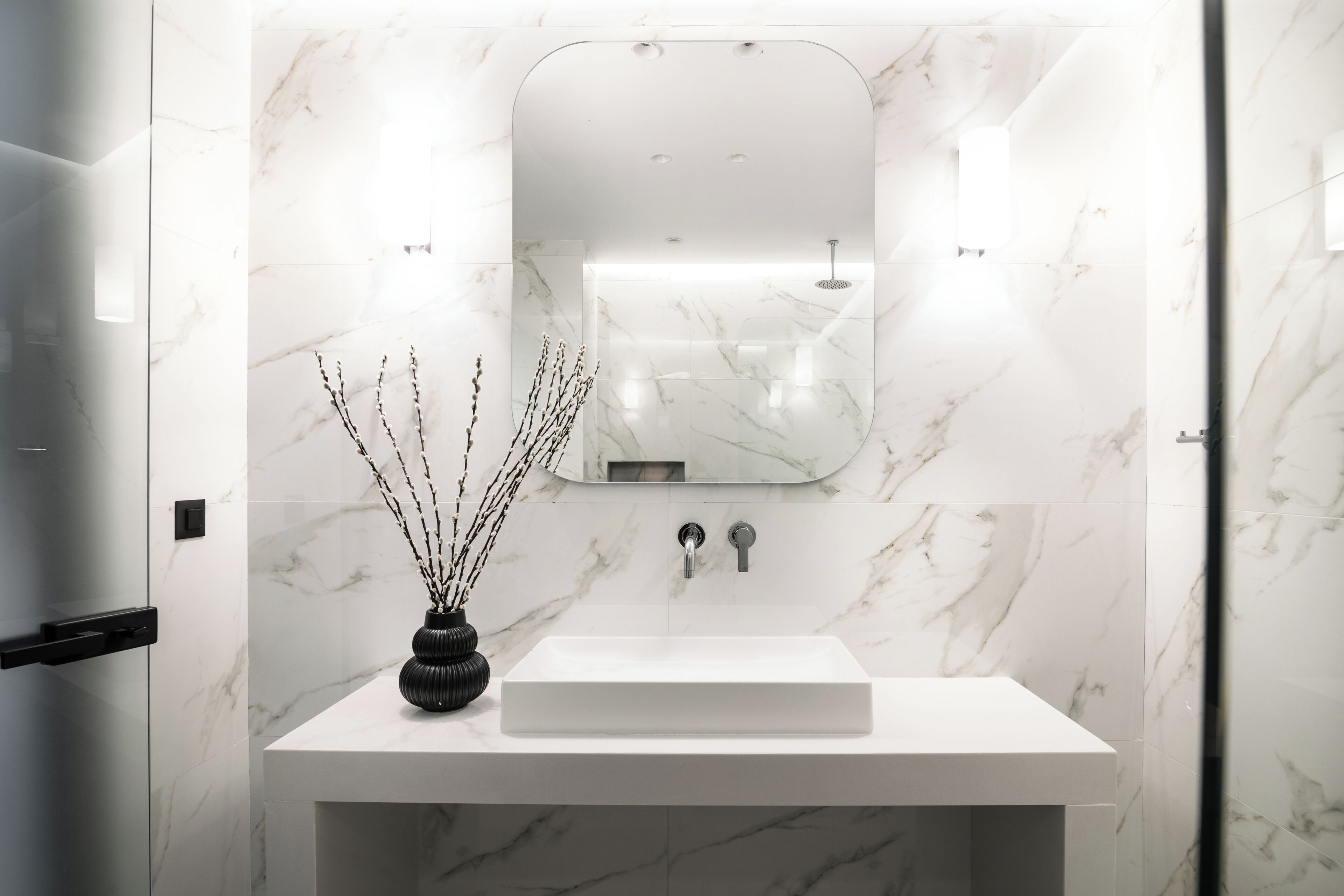 Un baño moderno con lavamanos de mármol blanco, un espejo y un jarrón negro con ramas.