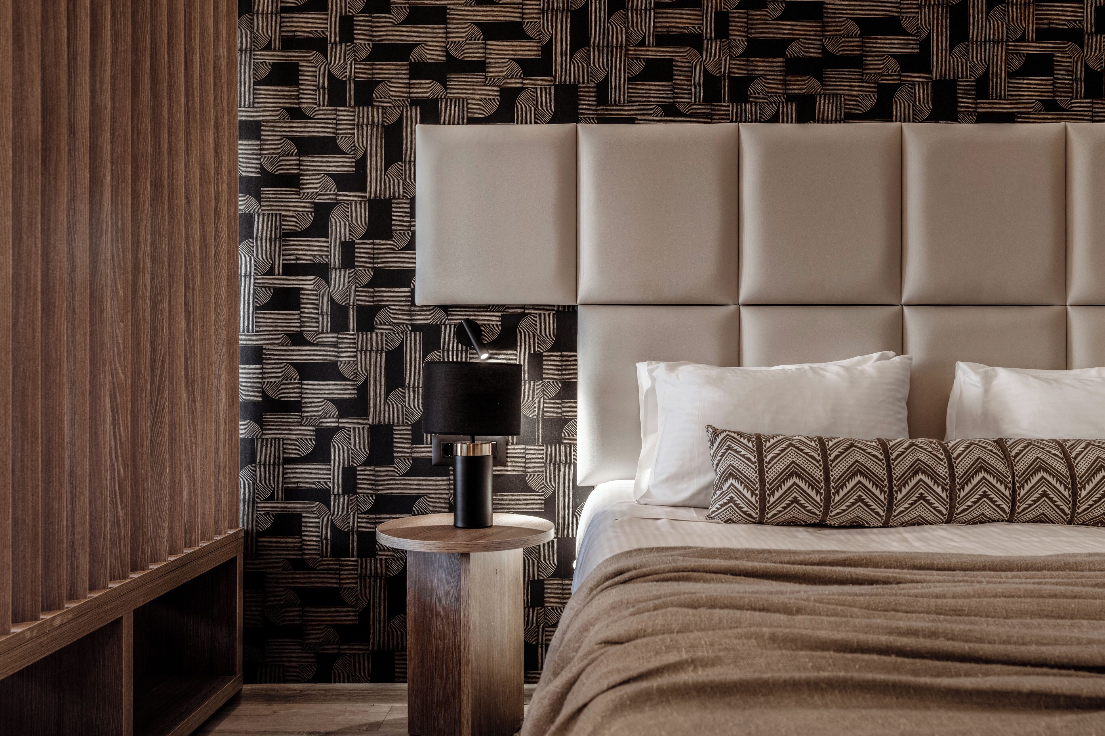 A bedroom with a tufted headboard, a round wooden nightstand, a black lamp, and patterned bedding.