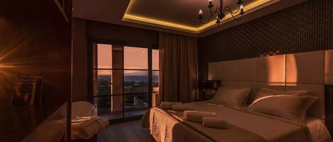 A modern hotel room with a large bed, a view of the cityscape through floor-to-ceiling windows, a chandelier, and a cozy seating area.
