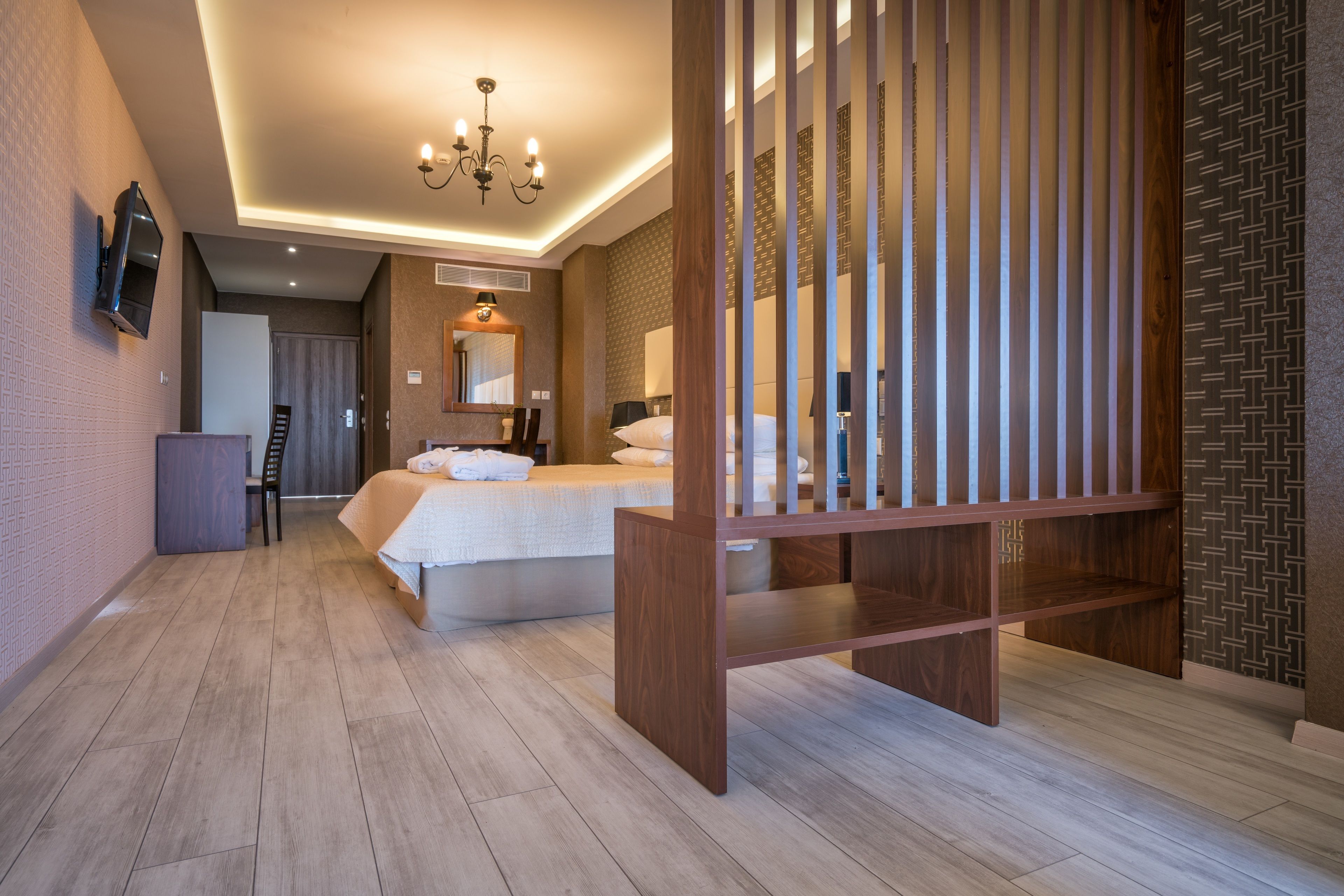 A hotel room with a bed, a wooden partition, a TV mounted on the wall, and a chandelier.