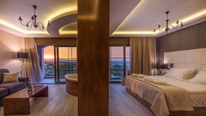 Una habitación de hotel con una cama grande, una zona de estar con un sofá y una mesita de café, una bañera y vistas a la puesta de sol a través de las ventanas.