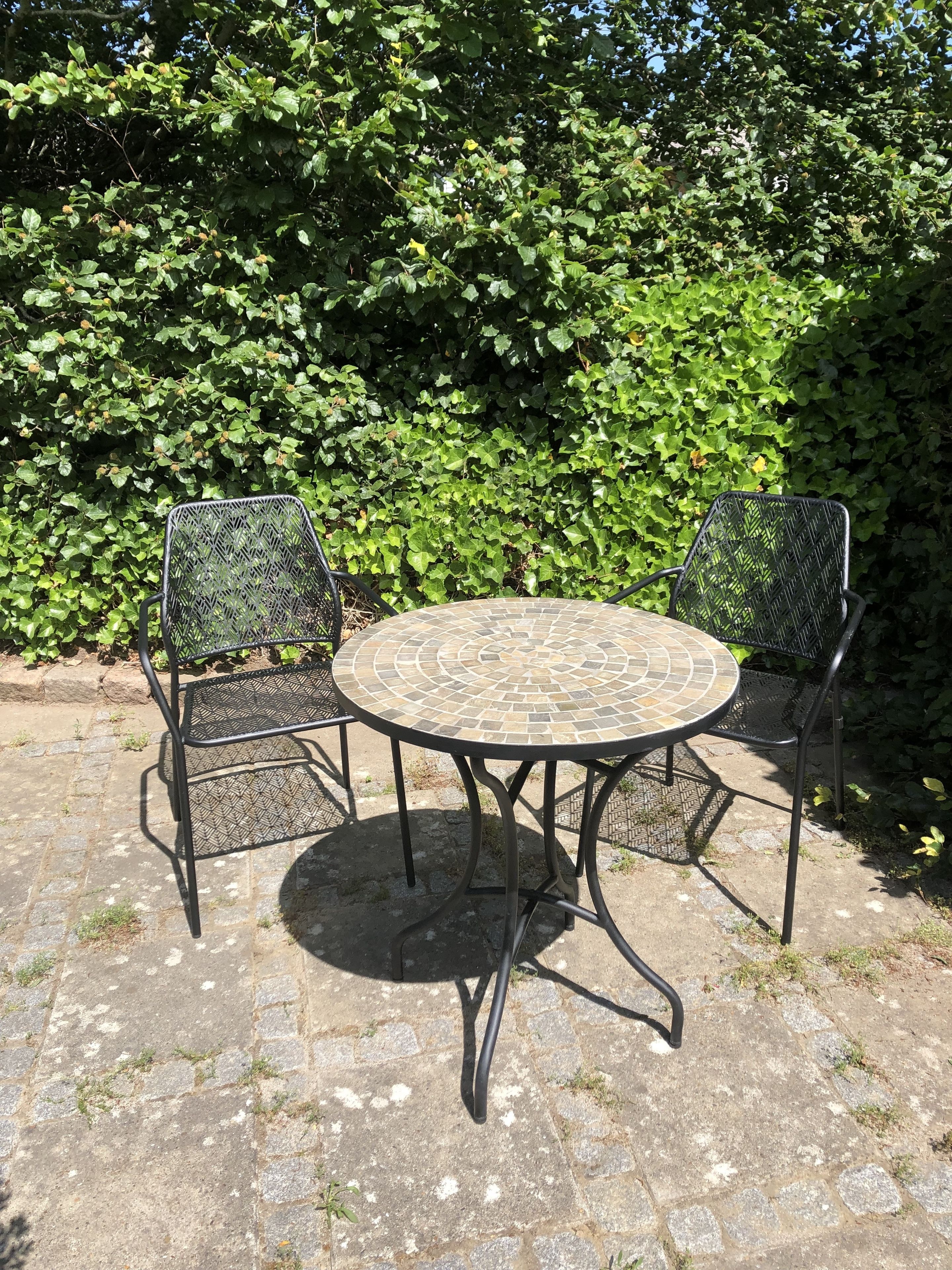 A round outdoor table with a mosaic top, surrounded by two metal chairs with a diamond pattern.